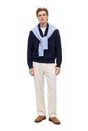 Homme portant un pull demi-zippé en cachemire bleu marine, avec un pull bleu clair en maille torsadée noué sur les épaules, un pantalon blanc cassé et des mocassins beige clair