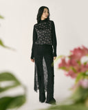 A woman in a black lace CashmereTech top and trousers beside pink blossom