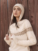 Woman in a cream Fair Isle cashmere jumper and matching ear-flap hat against a red wooden wall