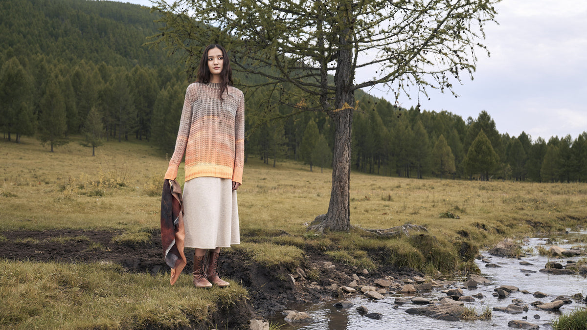 Woman by a stream on the Mongolian steppe wearing an orange-to-grey ombré cashmere knit over a cream skirt