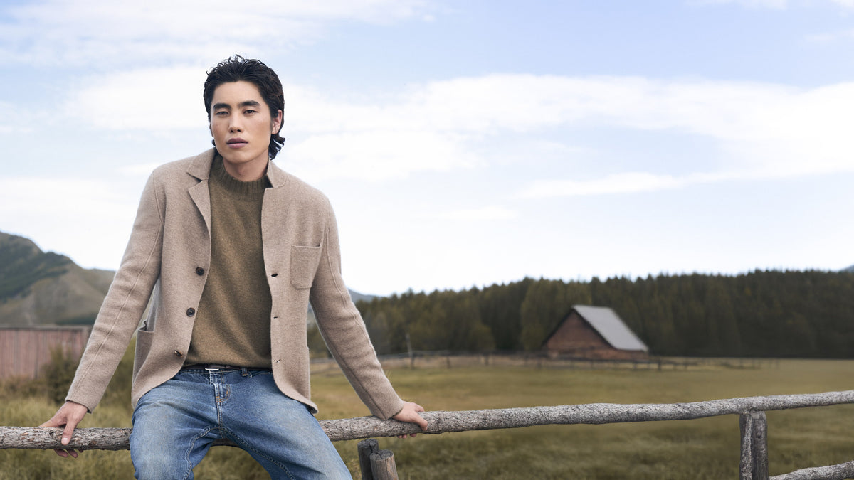 Man in a camel cashmere jacket and jumper sitting on a wooden fence in front of a barn