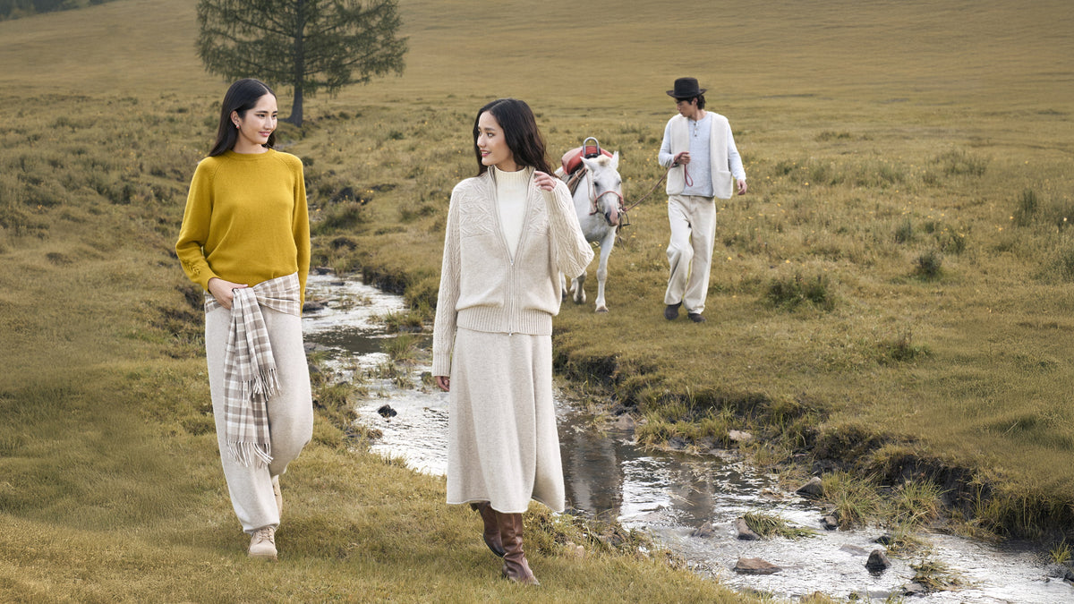 Two women in cashmere knits walking along a stream, a man leading a white horse behind them