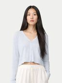 A woman with long dark hair wearing a light blue cropped V-neck cardigan and white pants, photographed against a plain light gray studio background.#color_soft-ice