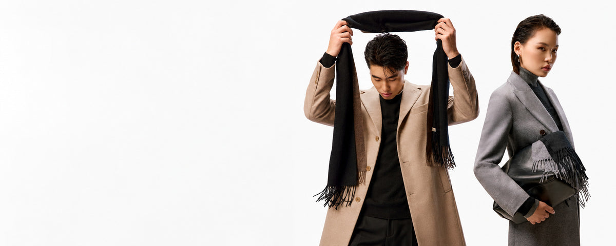 A man in a taupe cashmere coat lifts a black fringed scarf above his head, beside a woman in a grey coat holding a black leather bag and a fringed scarf