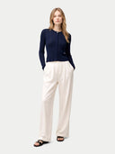 A woman modeling a navy blue long-sleeve knit top with vertical pleats and a small gold button detail at the collar, paired with off-white wide-leg trousers and black sandals, against a light gray studio background. #color_dark-indigo