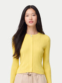 A woman with long dark hair wearing a bright yellow ribbed knit cardigan with a single button closure, paired with beige pants, against a light gray studio background. color_vivid-yellow