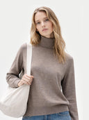 Woman wearing a brown turtleneck sweater holding a beige bag on a light gray background