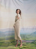 A woman in a cream ribbed cashmere top and midi skirt standing in profile against draped fabric printed with a Mongolian steppe landscape