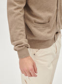 A man wearing a light brown knit V-neck cashmere cardigan over a white t-shirt, photographed against a solid light gray background. #color_taupe